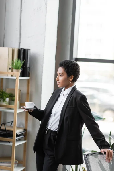 Молодая африканская американская бизнесвумен в черном костюме стоит с чашкой кофе и смотрит в сторону в офисе — стоковое фото