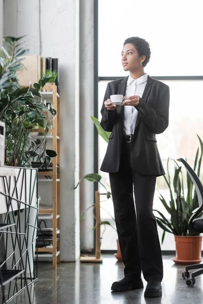 Piena lunghezza di donna d'affari afro-americana in abito nero in piedi con tazza di caffè vicino a piante in vaso verdi in ufficio moderno — Foto stock