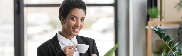 Freudige afrikanisch-amerikanische Geschäftsfrau im schwarzen Blazer mit Kaffeetasse und Blick in die Kamera im Büro, Banner — Stockfoto