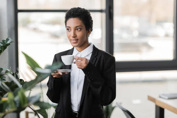 Sonriente mujer de negocios afroamericana en blazer negro de pie con taza de café y mirando hacia otro lado en la oficina - foto de stock