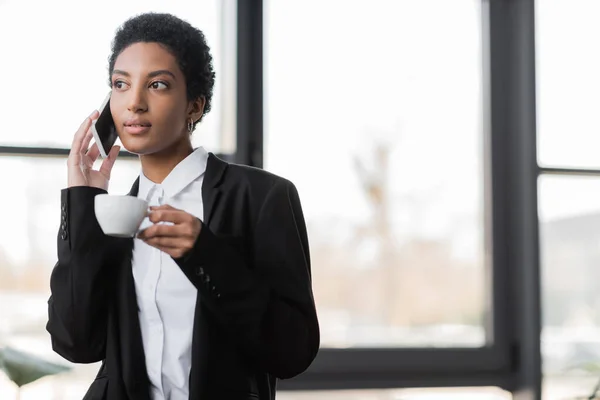 Elegante mujer de negocios afroamericana en chaqueta negra sosteniendo taza de café y hablando en el teléfono inteligente en la oficina - foto de stock