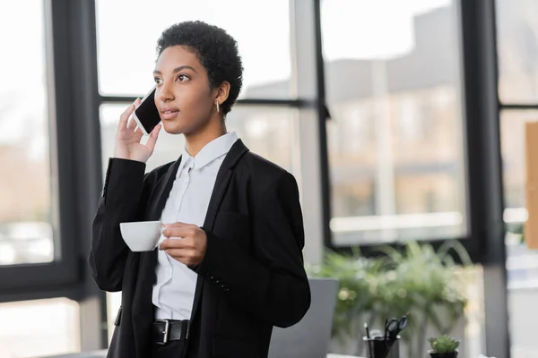 Mujer de negocios afroamericana positiva con taza de café mirando hacia otro lado durante la conversación en el teléfono móvil en la oficina - foto de stock