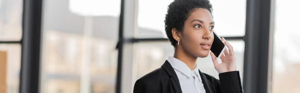 Joven mujer de negocios afroamericana mirando hacia otro lado mientras habla por teléfono celular en la oficina, pancarta - foto de stock