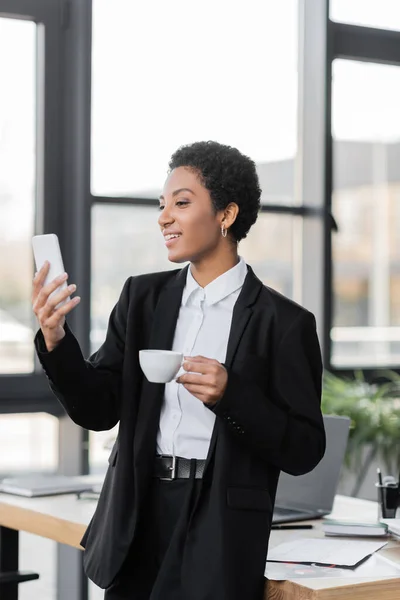 Allegra donna d'affari africana americana che tiene in mano una tazza di caffè e guarda lo smartphone in ufficio — Foto stock