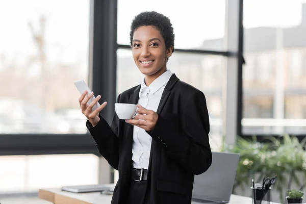 Agradecida mujer de negocios afroamericana con teléfono móvil y taza de café mirando a la cámara en la oficina - foto de stock