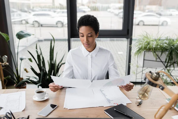 Afrikanisch-amerikanische Geschäftsfrau in weißer Bluse sitzt am Schreibtisch neben der Kaffeetasse und betrachtet Dokumente im modernen Büro — Stockfoto