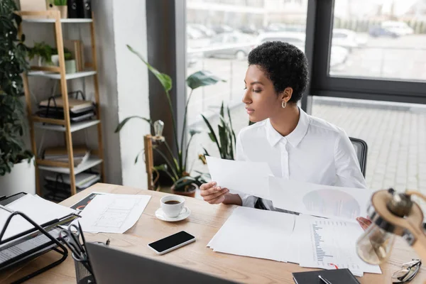 Elegante mujer de negocios afroamericana en blusa blanca trabajando con documentos cerca de taza de café y teléfono inteligente con pantalla en blanco en la oficina - foto de stock