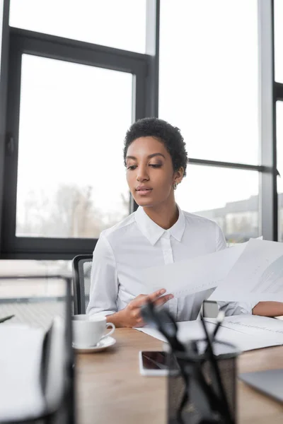 Elegante mujer de negocios afroamericana en blusa blanca sosteniendo documentos mientras está sentado cerca de la taza de café en la oficina - foto de stock