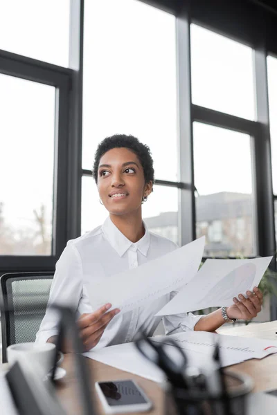 Alegre mujer de negocios afroamericana en blusa blanca sosteniendo documentos y mirando hacia otro lado en la oficina - foto de stock