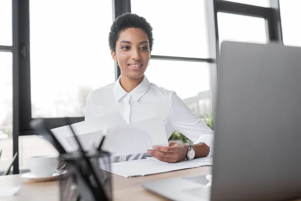 Heureuse femme d'affaires afro-américaine regardant ordinateur portable flou tout en travaillant avec des documents au bureau — Photo de stock