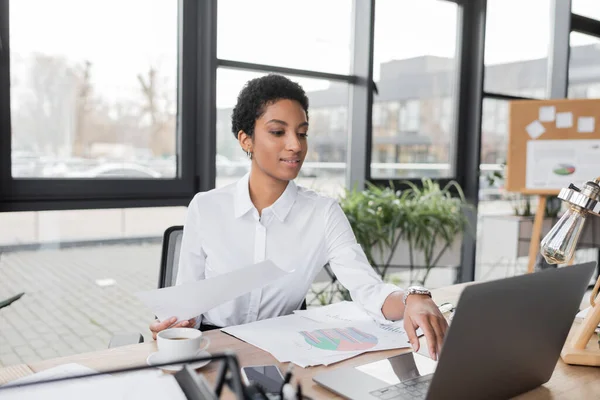 Усміхнена афроамериканська бізнес-леді використовує ноутбук біля документів на робочому столі в офісі — стокове фото