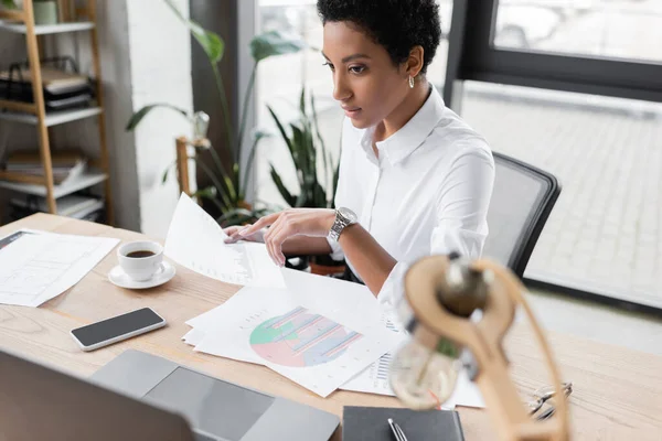 Mulher de negócios afro-americana segurando infográficos e olhando para laptop borrado enquanto trabalhava no escritório — Fotografia de Stock