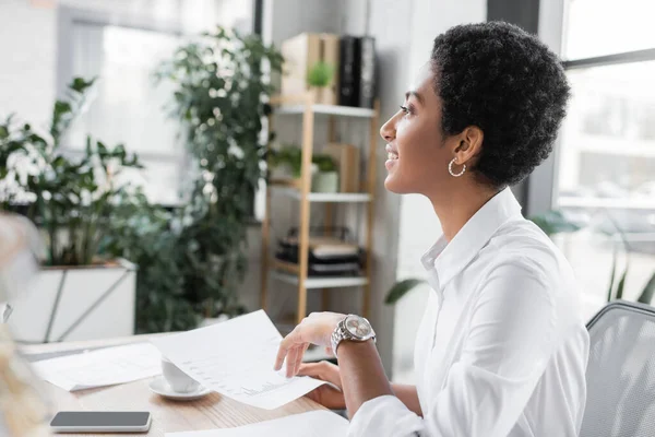 Vista lateral de la mujer de negocios afroamericana sonriendo y mirando hacia otro lado mientras trabaja con papeles en la oficina - foto de stock