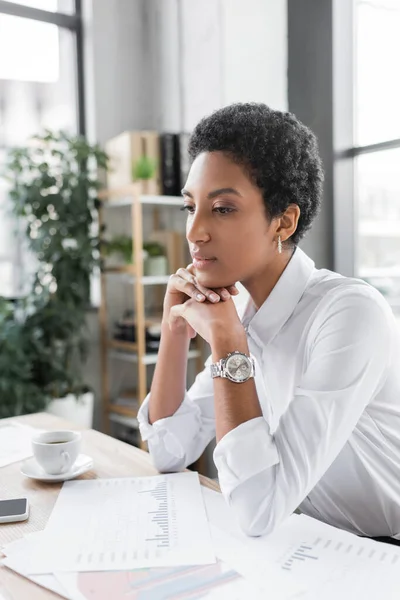 Розсудлива афроамериканська комерсантка сидить біля чашки з кавою і папірці з графіками на робочому столі. — стокове фото