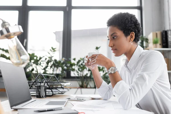 Side view of african american businesswoman holding cup of coffee and looking at laptop with blank screen in office — Stock Photo