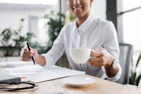 Cropped view of blurred african american businesswoman sitting at work desk and holding cup of coffee and pen in office — Stock Photo