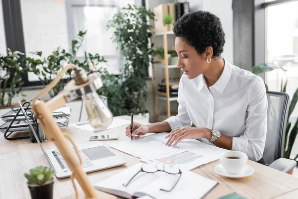 Молодая африканская американская бизнесвумен в белой блузке, пишущая на документах возле ноутбука и чашки кофе в современном офисе — стоковое фото