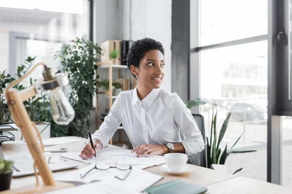 Feliz mulher de negócios afro-americana sentada com caneta perto de documentos e olhando para longe no escritório — Fotografia de Stock