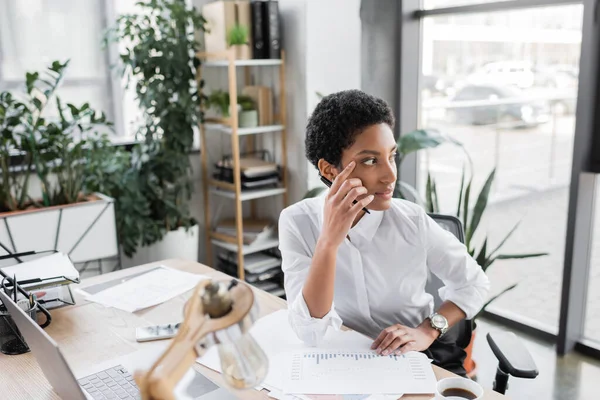 Pensativa mujer de negocios afroamericana sentada cerca de documentos y computadora portátil mientras mira hacia otro lado en la oficina - foto de stock