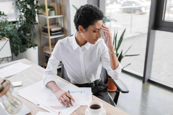 Tired african american businesswoman sitting near documents with charts and suffering from headache in office — Stock Photo