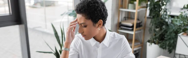 Agotada mujer de negocios afroamericana tocando la frente mientras sufre de dolor de cabeza en la oficina, pancarta - foto de stock