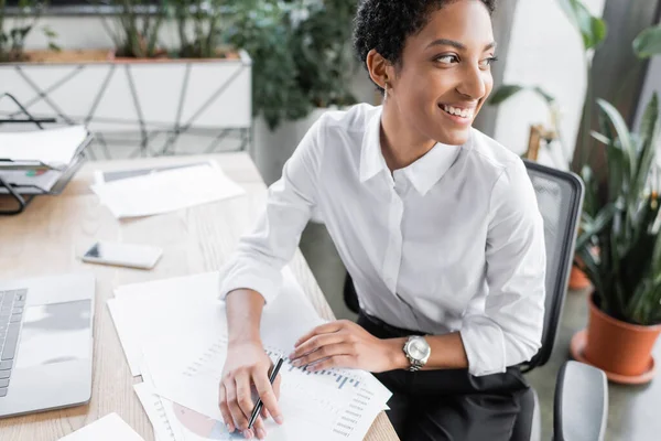 Alegre mujer de negocios afroamericana sentada cerca de documentos y gadgets y mirando hacia otro lado en la oficina - foto de stock
