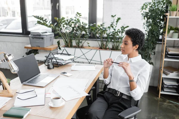 Africano americano empresária sentado com caneta no local de trabalho e olhando para laptop com tela em branco no escritório moderno — Fotografia de Stock