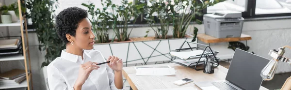 Стильна афроамериканська бізнесменка, що тримає ручку і дивиться на ноутбук з чистим екраном в сучасному офісі, банер — стокове фото