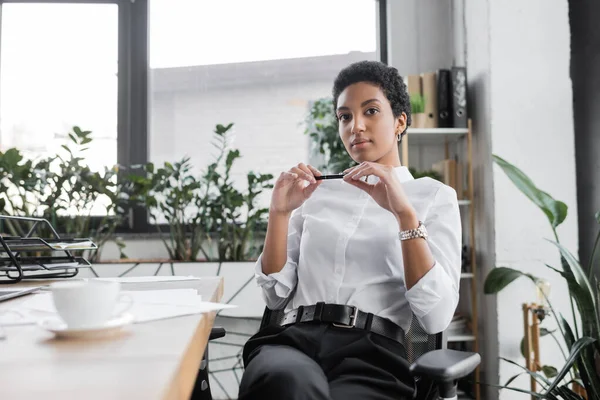Élégante femme d'affaires afro-américaine en chemisier blanc tenant stylo et regardant la caméra tout en étant assis dans un bureau moderne — Photo de stock