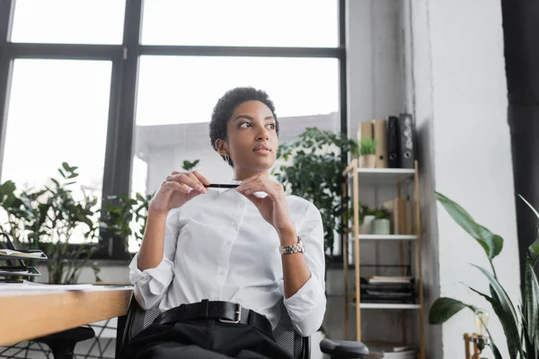 Pensativa mujer de negocios afroamericana en blusa blanca sosteniendo pluma y mirando hacia otro lado en la oficina - foto de stock