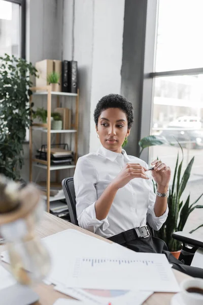 Nachdenkliche afrikanisch-amerikanische Geschäftsfrau in weißer Bluse hält Stift in der Hand, während sie neben Dokumenten im Büro sitzt — Stockfoto