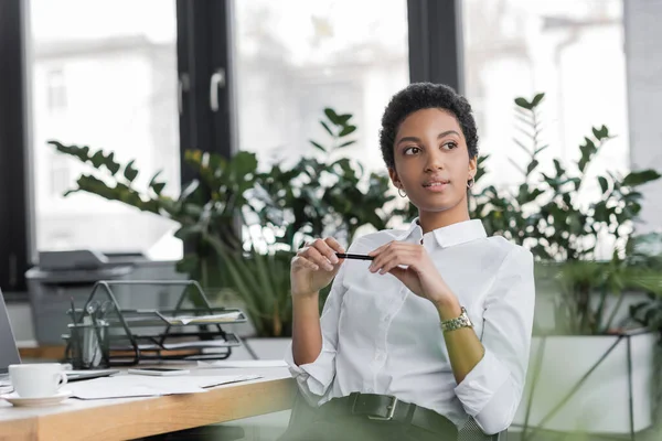 Mujer de negocios afroamericana positiva y soñadora sosteniendo la pluma y mirando hacia otro lado mientras está sentada en la oficina - foto de stock