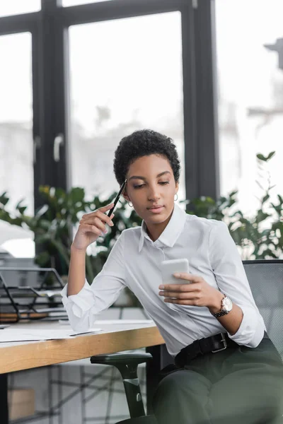 Вдумчивая американская бизнесвумен в белой блузке, держащая ручку и глядя на смартфон в офисе — стоковое фото