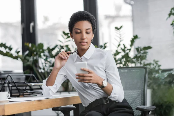 African american businesswoman holding pen and using cellphone while sitting at workplace in office — Stock Photo