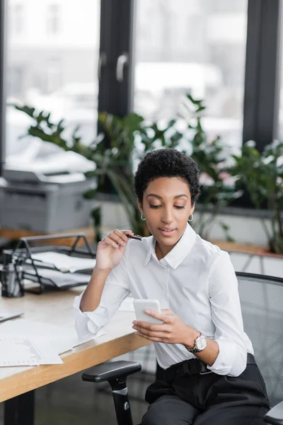 Jeune femme d'affaires afro-américaine en chemisier blanc tenant stylo et en utilisant un ordinateur portable tout en étant assis au bureau — Photo de stock