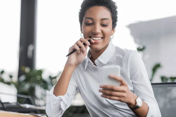 Gioiosa donna d'affari africana americana in camicetta bianca che tiene la penna e guarda il telefono cellulare in ufficio — Foto stock