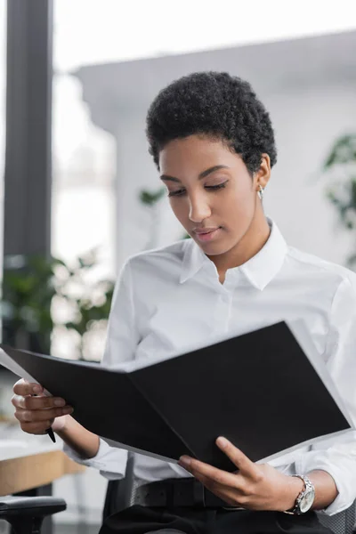 Jeune femme d'affaires afro-américaine en chemisier blanc lisant des documents dans le dossier au bureau — Photo de stock