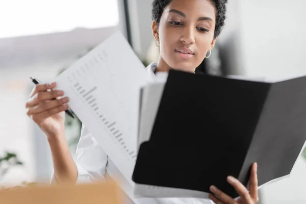Молода афроамериканська бізнес-леді тримає теку і дивиться на документи з інфографікою в офісі — стокове фото