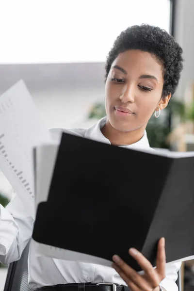 Африканская американская бизнесвумен держит папку и просматривает документы с диаграммами в офисе — стоковое фото