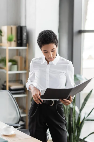 Elegante mujer de negocios afroamericana en blusa blanca y pantalones negros de pie con carpeta en la oficina - foto de stock