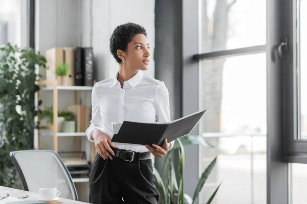 Афроамериканська бізнесменка в стильному офіційному одязі тримає папку з документами і дивиться у вікно в сучасному офісі. — стокове фото