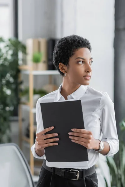 Pensativa mujer de negocios afroamericana sosteniendo carpeta con documentos y mirando hacia otro lado en la oficina - foto de stock