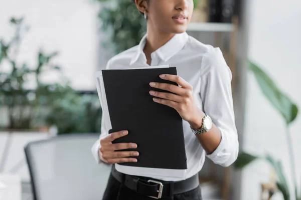 Vista recortada de la mujer de negocios afroamericana en blusa blanca que sostiene la carpeta con documentos en la oficina - foto de stock