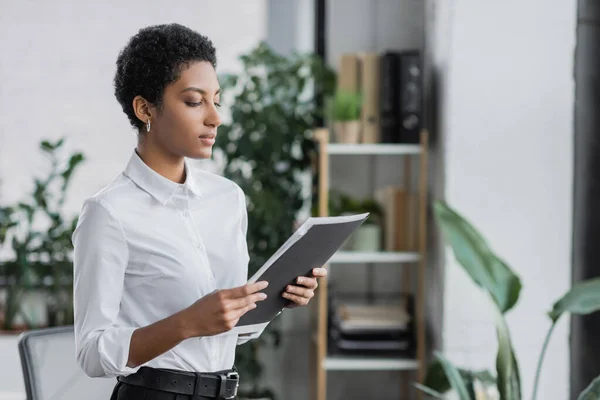 Junge afrikanisch-amerikanische Geschäftsfrau in weißer Bluse schaut auf Ordner mit Dokumenten im Büro — Stockfoto