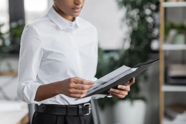 Vista parcial del joven gerente afroamericano en blusa blanca sosteniendo carpeta con documento mientras está de pie en la oficina - foto de stock