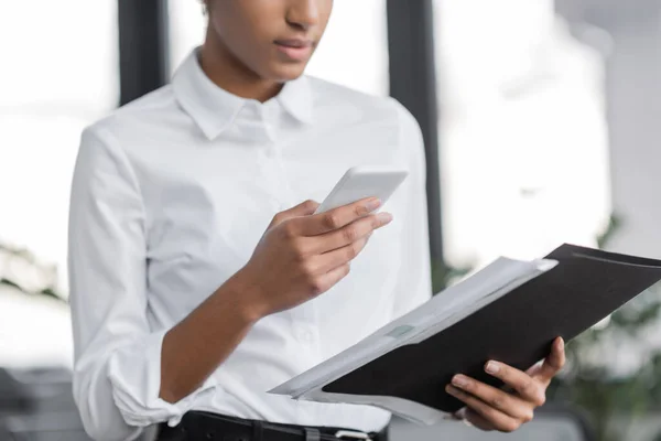Vista recortada de la mujer de negocios afroamericana en blusa blanca que sostiene la carpeta y mensajería en el teléfono inteligente en la oficina - foto de stock