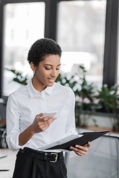 Усміхнена афроамериканська бізнес-леді тримає смартфон і дивиться на папку з документами в офісі — стокове фото