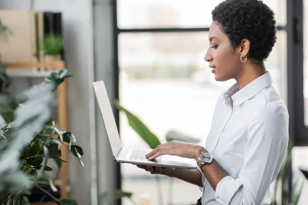 Бічний вид на молоду афроамериканську комерсантку, що стоїть в офісі і використовує ноутбук з чистим екраном — Stock Photo