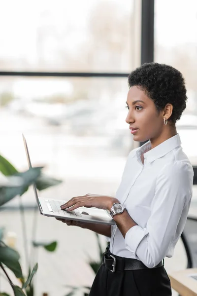 Side view of dreamy african american businesswoman in white shirt holding laptop and looking away in office — Stock Photo