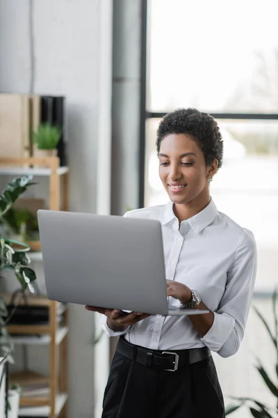 Cheerful african american businesswoman using laptop while standing in modern office — Stock Photo